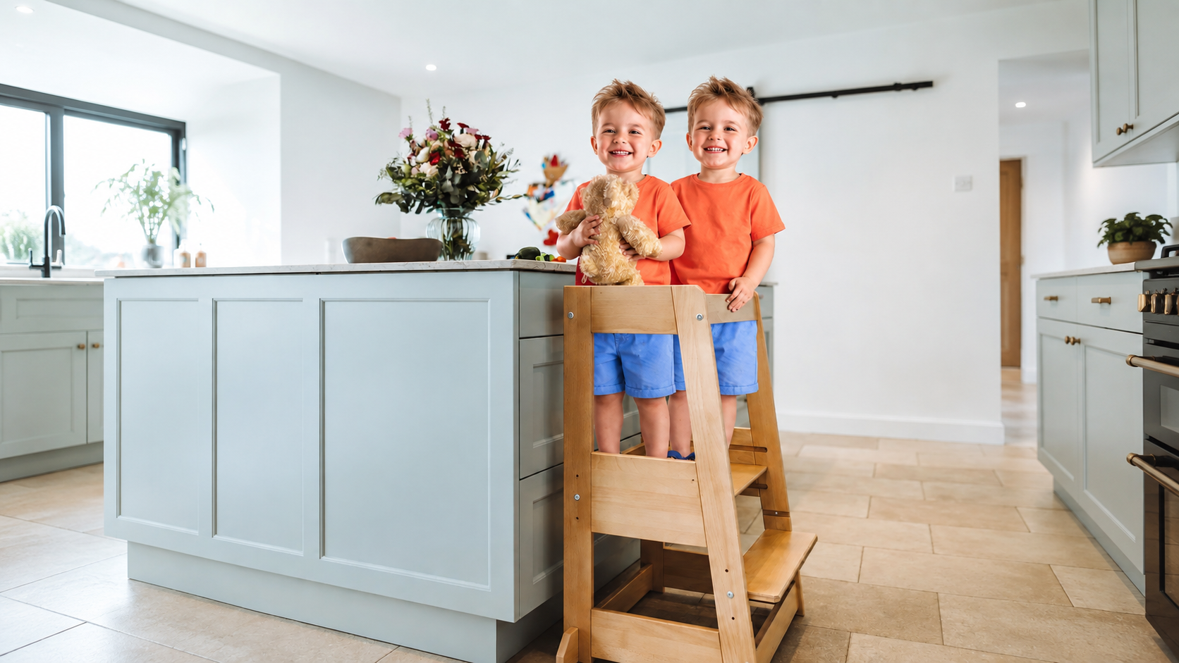 Zwei fröhliche Kinder stehen gemeinsam auf einem Holz-Lernturm in der Küche – stimmungsvolles Bild für Geschenkideen zur Geburt von Zwillingen und personalisierte Zwillingsgeschenke.