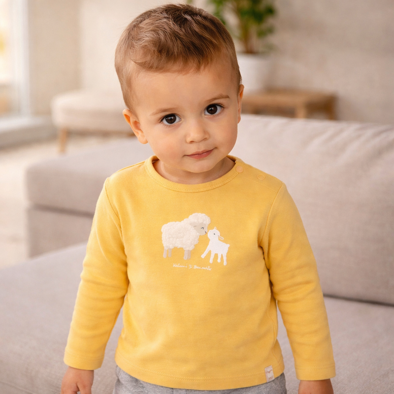 Kleiner Junge trägt ein gelbes langärmeliges Shirt mit Schaf Motiv und steht in einem hellen Wohnzimmer